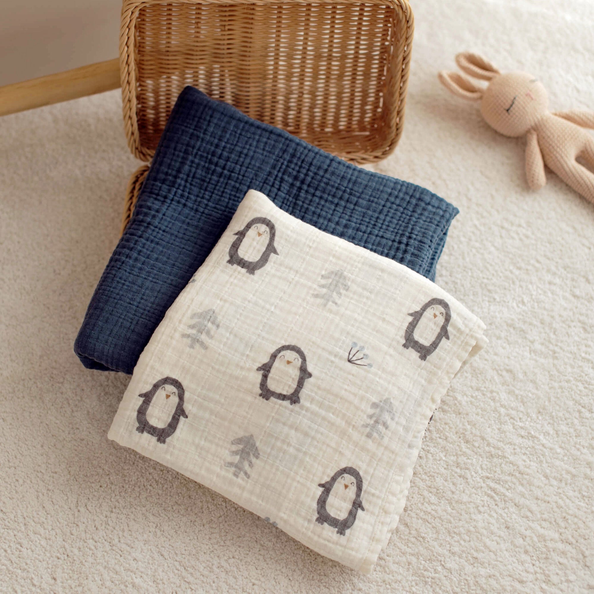 Penguin swaddle and navy blanket folded in a basket with toy nearby