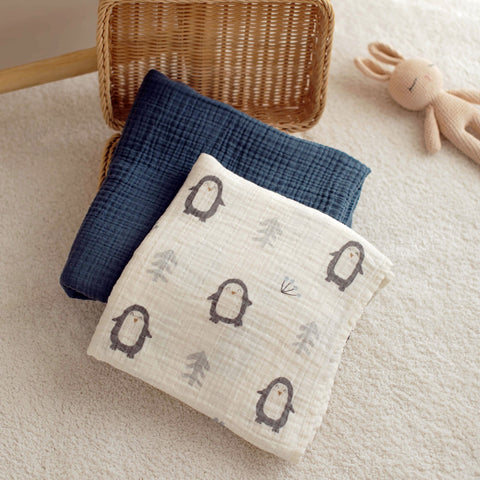 Penguin swaddle and navy blanket folded in a basket with toy nearby