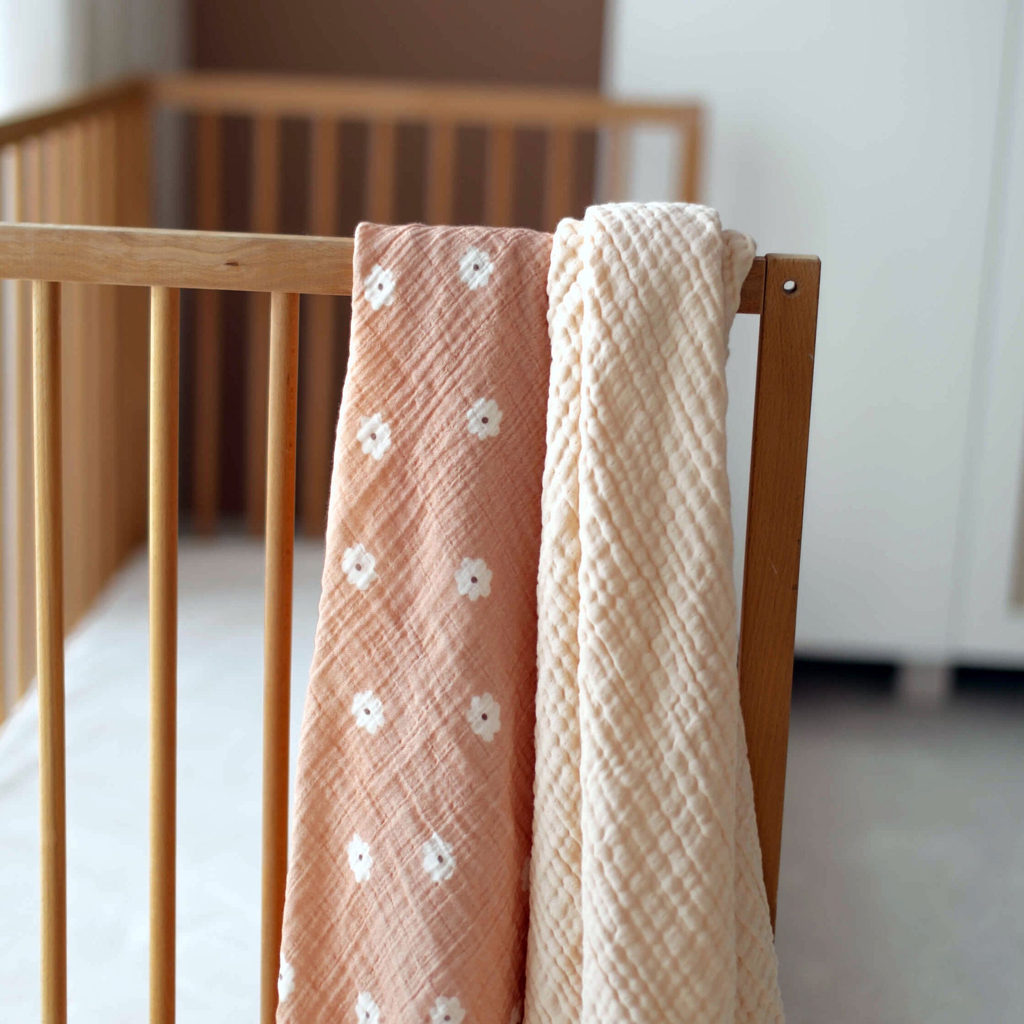 Muslin swaddle and 4-ply cream blanket hanging over wooden crib rail
